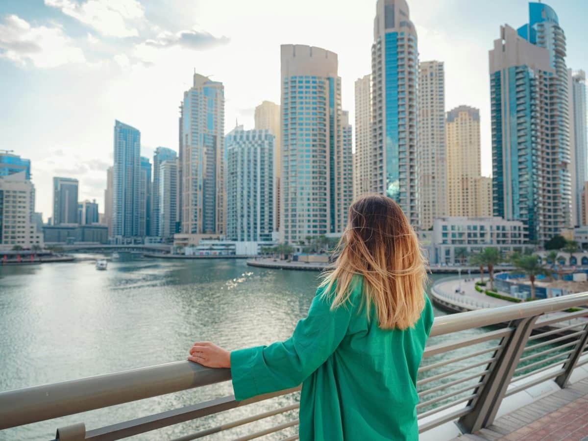 Une personne portant un manteau vert regarde l'horizon d'une ville au bord de l'eau, debout sur un pont avec des gratte-ciels modernes en arrière-plan sous un ciel partiellement nuageux.
