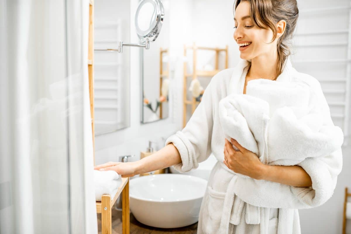 Femme en peignoir blanc tenant des serviettes et souriant dans une salle de bain moderne avec un lavabo et un miroir en arrière plan.