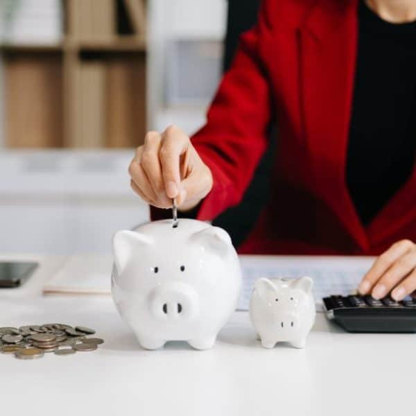 Une personne portant un blazer rouge met une pièce dans une tirelire sur un bureau avec des pièces de monnaie, une tirelire plus petite, une calculatrice et un ordinateur portable.