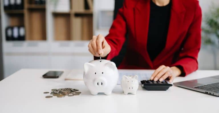 Une personne portant un blazer rouge met une pièce dans une tirelire sur un bureau avec des pièces de monnaie, une tirelire plus petite, une calculatrice et un ordinateur portable.