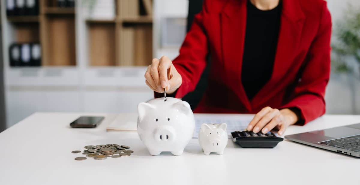 Une personne portant un blazer rouge met une pièce dans une tirelire sur un bureau avec des pièces de monnaie, une tirelire plus petite, une calculatrice et un ordinateur portable.