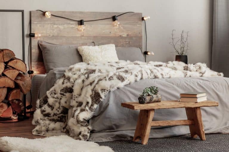 Chambre cosy avec tête de lit en bois rustique, guirlandes lumineuses et couverture en fourrure moelleuse. Un banc en bois accueille des livres et une petite plante. Des bûches sont soigneusement empilées à côté du lit.