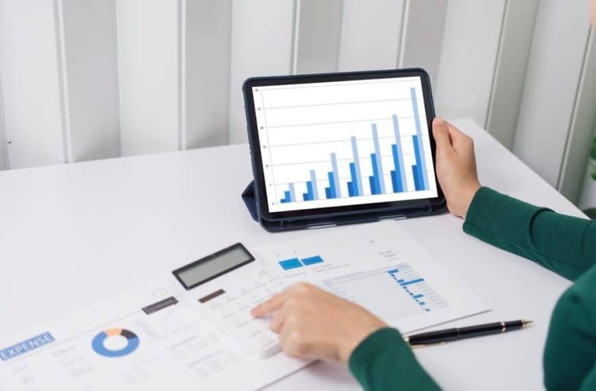 Personne en chemise verte examinant un graphique à barres sur une tablette au bureau avec des documents, une calculatrice et un stylo.