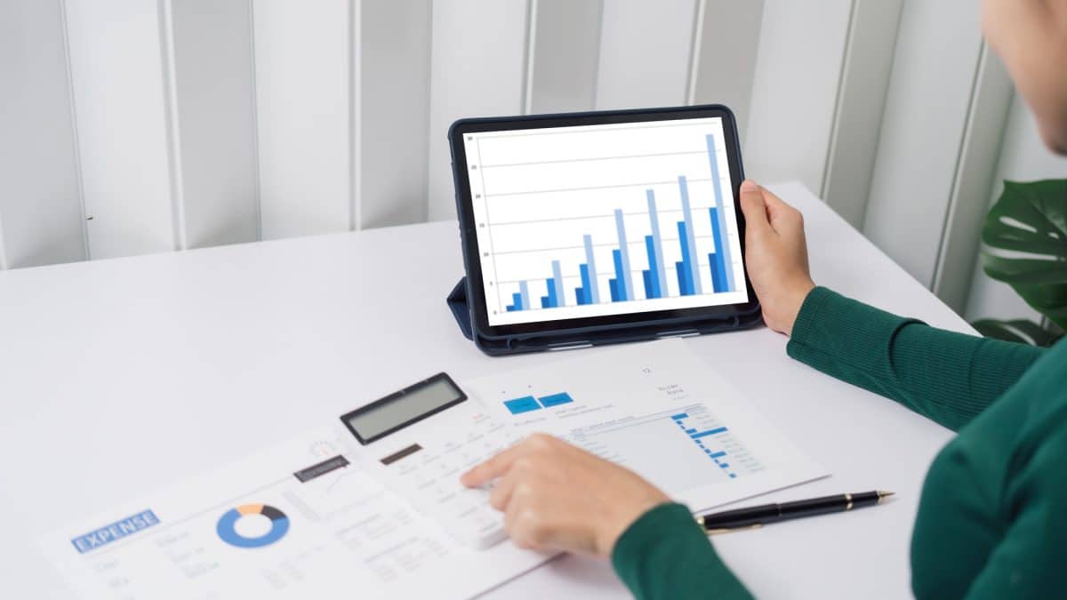Personne en chemise verte examinant un graphique à barres sur une tablette au bureau avec des documents, une calculatrice et un stylo.
