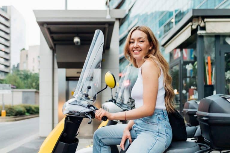 Une femme aux cheveux longs est assise sur un scooter jaune dans une zone urbaine et sourit à l'appareil photo. Elle porte un débardeur blanc et un jean bleu.