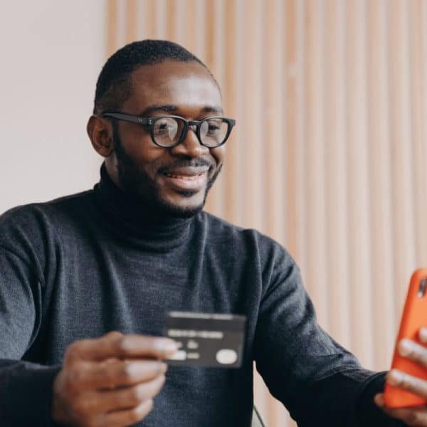 Un homme portant des lunettes et un col roulé sombre tient une carte de crédit dans une main et un smartphone dans l'autre, souriant en regardant le téléphone.
