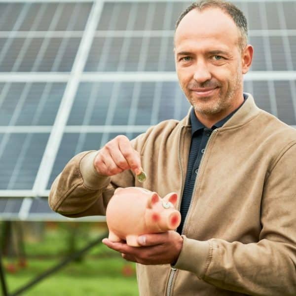 Un homme vêtu d'une veste marron met une pièce de monnaie dans une tirelire rose alors qu'il se tient devant des panneaux solaires.