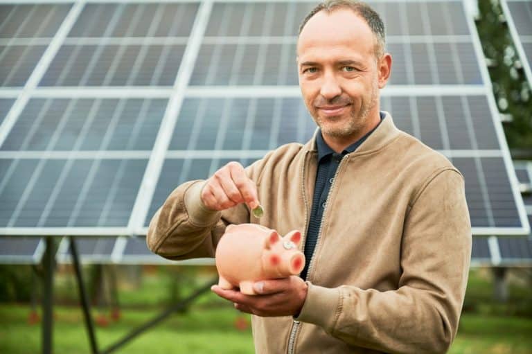 Un homme vêtu d'une veste marron met une pièce de monnaie dans une tirelire rose alors qu'il se tient devant des panneaux solaires.