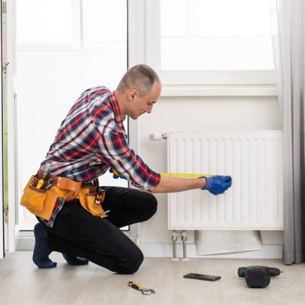 Un homme portant des gants de travail et une ceinture à outils est agenouillé sur le sol, mesurant un radiateur blanc avec un ruban à mesurer dans une pièce lumineuse.