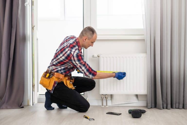 Un homme portant des gants de travail et une ceinture à outils est agenouillé sur le sol, mesurant un radiateur blanc avec un ruban à mesurer dans une pièce lumineuse.