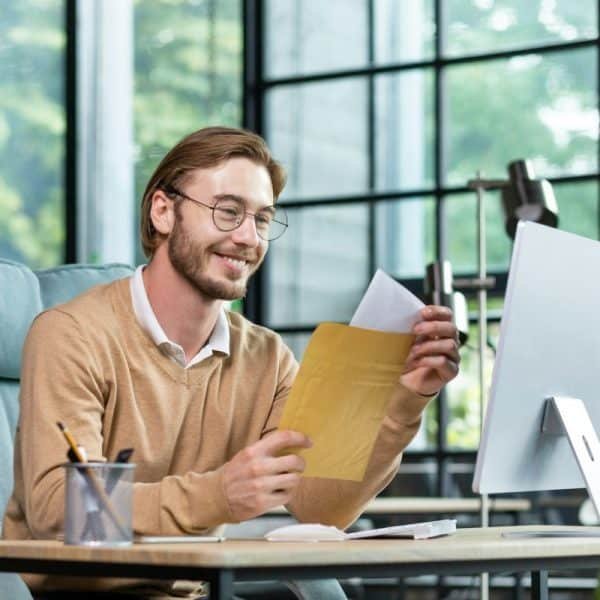 Un homme portant des lunettes et un pull beige est assis à un bureau devant un ordinateur, ouvrant une enveloppe avec un sourire. Un pot à crayons et divers articles de bureau sont posés sur le bureau.