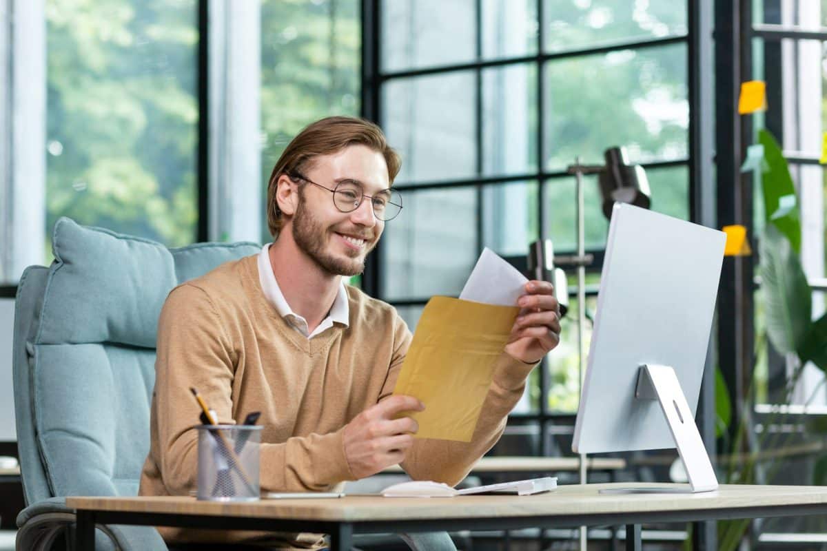 Un homme portant des lunettes et un pull beige est assis à un bureau devant un ordinateur, ouvrant une enveloppe avec un sourire. Un pot à crayons et divers articles de bureau sont posés sur le bureau.